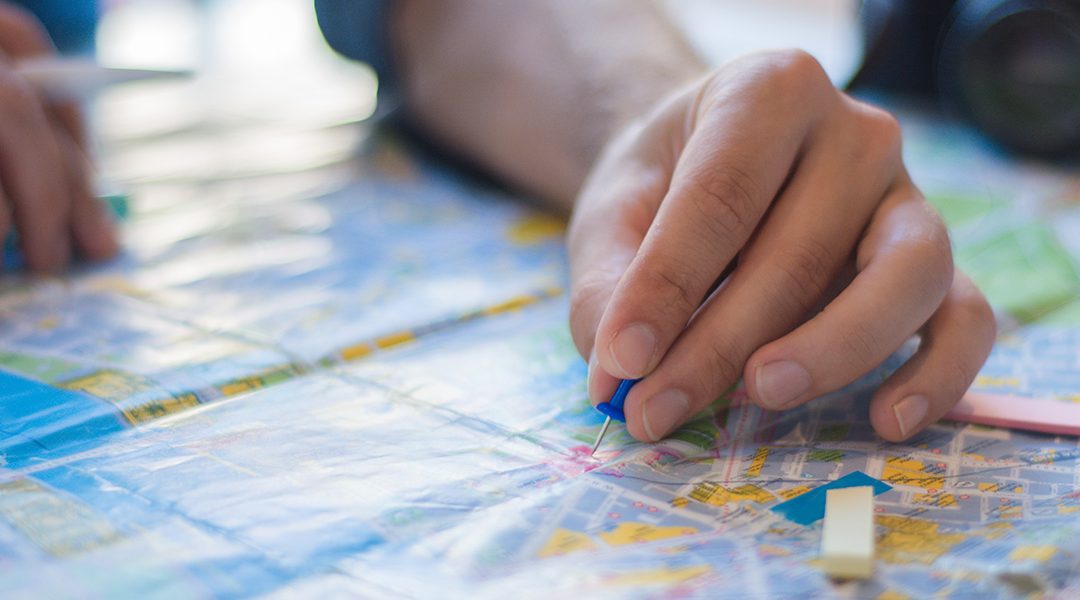 Close-up of hands marking a city map with a blue pencil and sticky notes on a travel planning table.