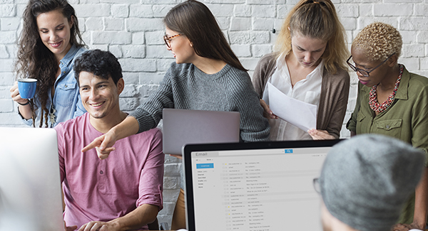 Diverse team of five coworkers collaborating around laptops in a modern office, with one person pointing at a screen while others review papers.