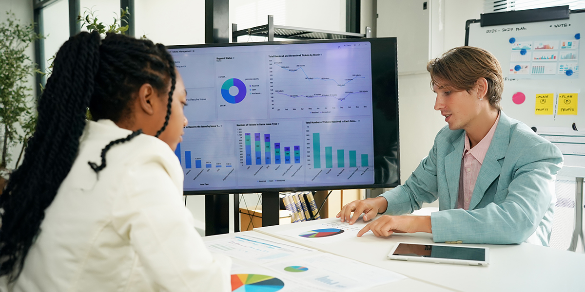 Two professionals discuss data charts displayed on a large monitor, with printed graphs on the table's surface.