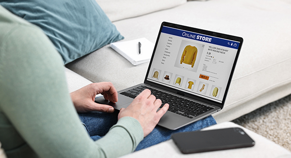 Person browsing an online clothing store on a laptop, viewing a product page with a yellow jacket and related items on screen near a sofa and phone.