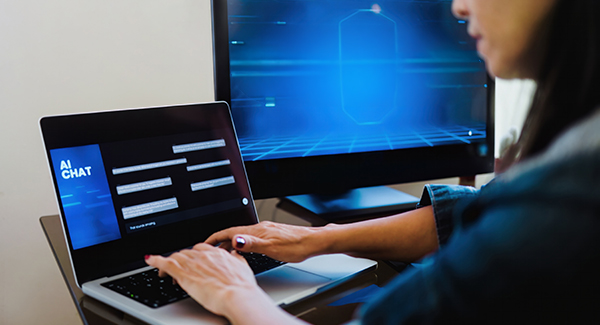 Person typing on a laptop showing an AI chat interface; blue digital monitor in the background.